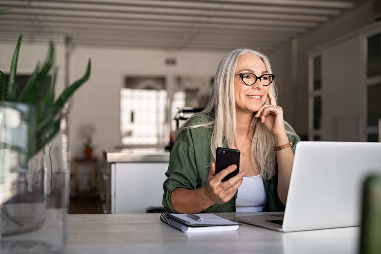 Happy senior woman holding smartphone and laptop daydreaming while looking away